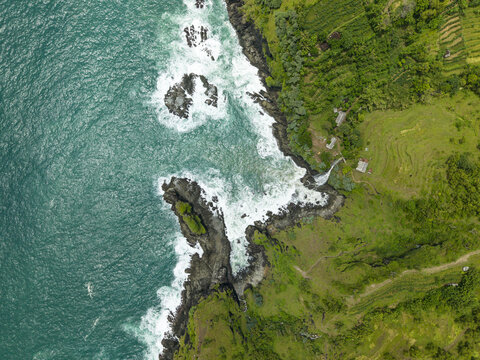 Aerial view of Banyutibo Water Fall cascading over dark cliffs into the turquoise ocean waves and lush green terraced fields Girisubo, Special Region of Yogyakarta, Indonesia.