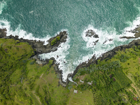 Aerial view of Banyutibo Water Fall cascading from lush green cliffs into the turquoise ocean waves in Girisubo, Special Region of Yog, Indonesia.