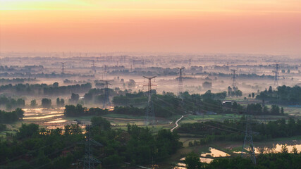 This scenic dawn rural landscape features mist-shrouded farmlands, dense woodlands and high voltage transmission towers under a soft orange-pink sky. © tai