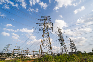 Multiple steel lattice transmission towers and power lines stand in a grassy field under a blue cloudy sky, part of electrical power infrastructure. © tai