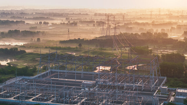 Aerial view of a high voltage substation and transmission towers at misty dawn, with rural wetlands and open fields under warm morning sunlight.