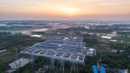 This aerial shot shows a large high voltage power substation at dawn, surrounded by rural wetlands and woodlands under a sunrise sky. © tai