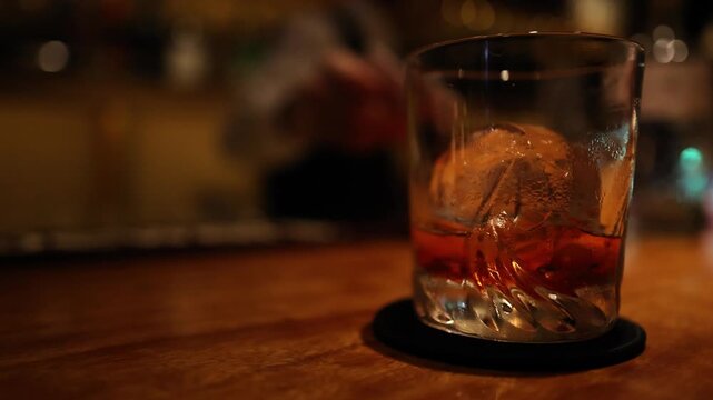 A whiskey glass with a spherical ice cube sits on a dark coaster on a wooden counter, with a blurred bartender in background.