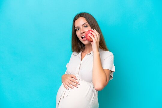 Young caucasian woman isolated on blue background pregnant and holding an apple and eating it