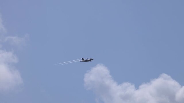 Modern military stealth jet flying through blue sky with vapor trails and white clouds.