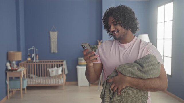 Man cradles a wrapped baby with one arm while checking a smartphone in a baby room building, standing near a crib and lamp; quiet tenderness.