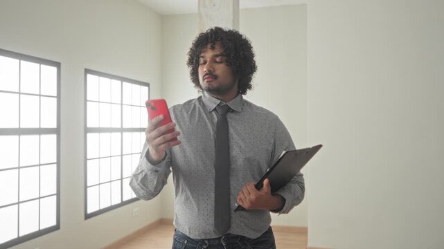 Young indian man right hand holding red smartphone and left arm clutching clipboard inside empty building; focused professionalism.