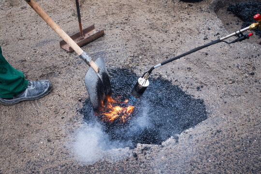 Road workers repair a pothole on a street by pouring hot asphalt from a bag and smoothing it with a torch and shovel.