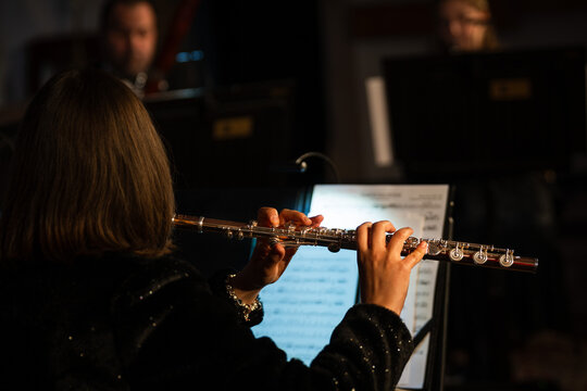 Rear view of a female flutist in a black sequined top playing a silver transverse flute during an orchestra performance, with sheet music visible.