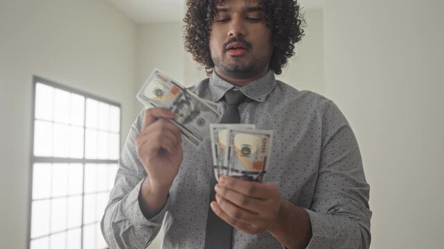 Man counts dollars and thumbs through a stack of cash, holding bills near chest in a building with tall window light; wealth planning confidence.