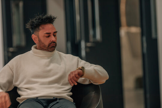 Young man checking his watch while sitting in a modern office, expression of impatience and stress