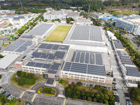 Aerial view of large industrial factory buildings with solar panels on rooftops and parking lots surrounded by green trees in Dongguan, Guangdong Province, China.