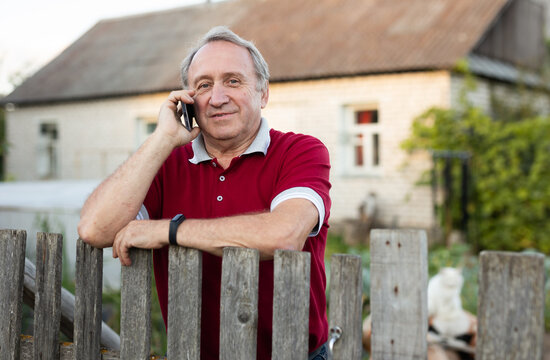 Elderly farmer talking on mobile phone at the fence of his farm on summer evening