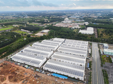 Aerial view of large industrial warehouses with solar panels on roofs surrounded by lush green trees and construction sites under an overcast sky An Phuoc, Dong Nai, Vietnam.