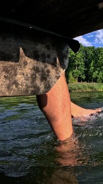 Close up of a man's legs dangling from a wooden pier, kicking and splashing in the cool lake water on a beautiful sunny day, enjoying a relaxing and carefree summer vacation activity