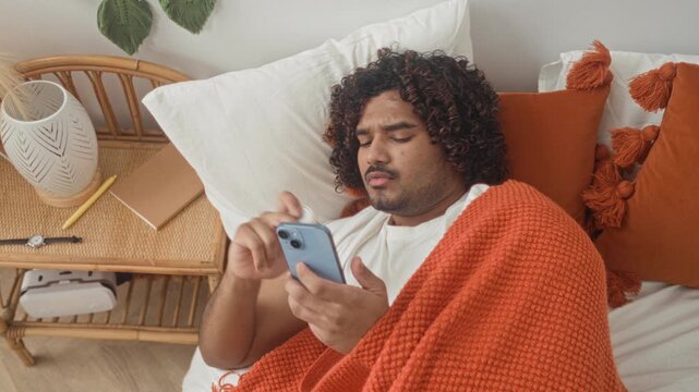 Man indian young lying in bed under an orange blanket holding smartphone and pill bottle, reading label by bedside table in a building; concern selfcare.