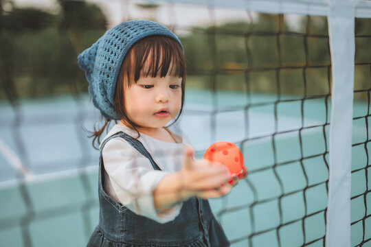 Cute Asian toddler girl holding an orange ball on a pickleball court at sunset. Happy child in denim outfit learning sports, promoting active lifestyle, wellness, and childhood development