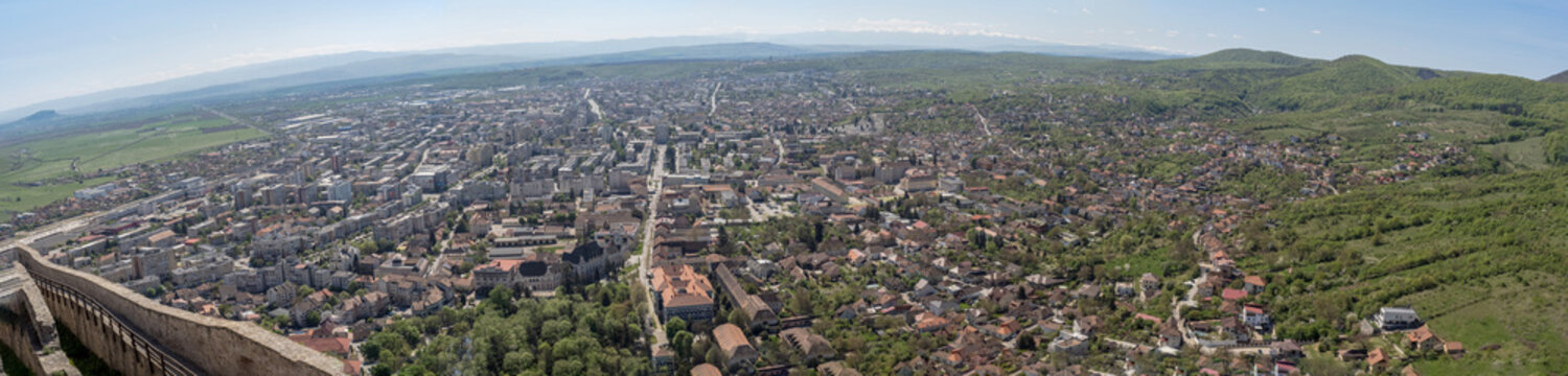 Wide aerial panorama of Deva city, Romania, taken from the Citadel hill, showcasing the urban landscape with a mix of historical and modern architecture, residential areas, and green hills