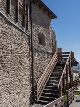 Ancient stone ruins and massive walls of the medieval Deva Citadel (Cetatea Devei) in Romania, featuring a tourist waving from a defensive platform and modern glass-and-wood structures