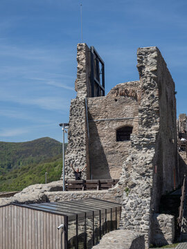 Ancient stone ruins and massive walls of the medieval Deva Citadel (Cetatea Devei) in Romania, featuring a tourist waving from a defensive platform and modern glass-and-wood structures