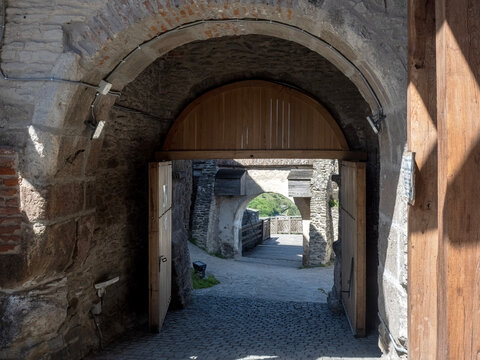 Exterior view of the main gate and massive stone fortifications of the medieval Deva Citadel in Romania, featuring a defensive tower with a red conical roof, a cobblestone path, and wooden stairs