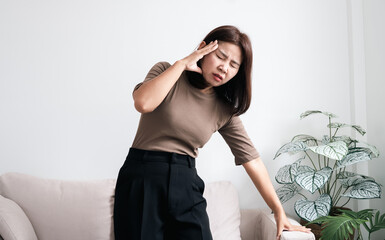 Naklejka premium Asian woman feeling dizzy and faint, holding her head and leaning on sofa for support. Concept of vertigo, hypertension, morning sickness, or low blood pressure. Healthcare and medical theme