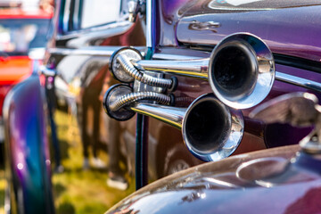 Naklejka premium A close-up shot of shiny chrome dual air horns mounted on the side of a purple, custom vintage car, highlighting metal reflections and intricate details in bright daylight.