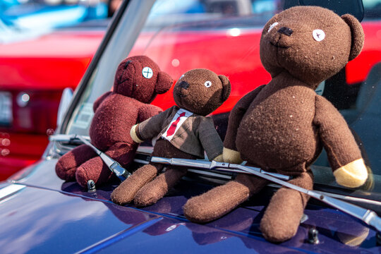 Three brown plush teddy bears sitting on the purple hood of a vintage car below the windshield, including one in a suit and tie, with a white windshield frame and a red car in the background, in brigh