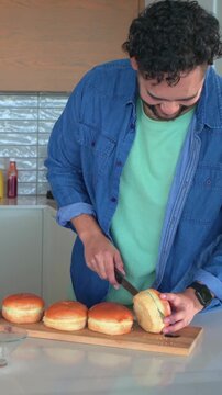 Slicing adult male in denim shirt at kitchen counter, cutting buns on wooden board with knife