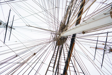 Obraz premium The tall masts and complex rigging of a ship taken from a low angle. The lines stretch in all directions against a light sky. The ship is moored and not moving.