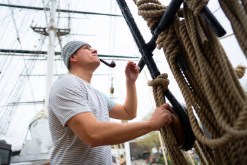 A sailor stands on a tall ship, adjusting ropes and looking up. He wears a striped shirt and a beanie while holding a pipe. The ship's masts are visible in the background. © Videophilia