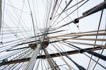 Fototapeta premium The view shows the intricate arrangement of ropes and masts on a sailing ship as seen from below. The sky appears cloudy, creating a muted atmosphere.