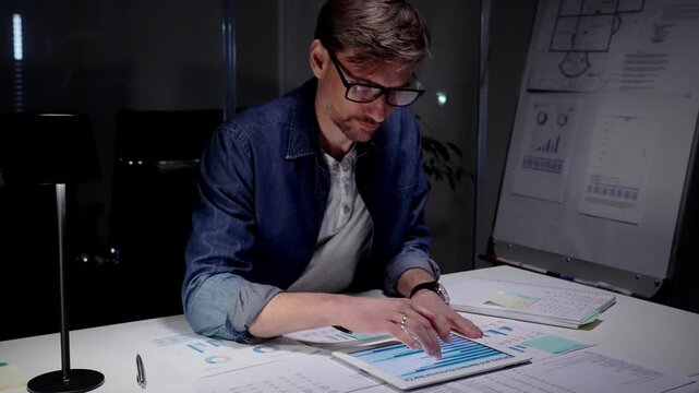 Professional man working late in a dark office, analyzing business data reports and using a digital tablet, managing overtime and workload under focused concentration
