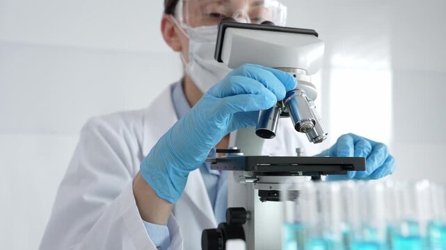 Female scientist wearing protective equipment checking microscope, analyzing blue test tubes in modern laboratory research setting. Medicine, healthcare and science concept