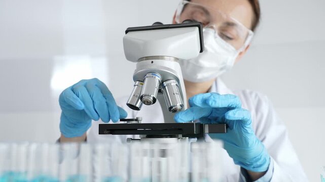 Female scientist wearing protective equipment checking microscope, analyzing blue test tubes in modern laboratory research setting. Medicine, healthcare and science concept
