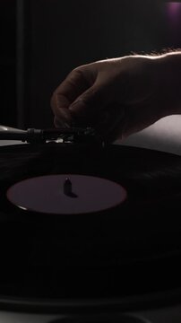 Vertical video human hand lowering tonearm onto a spinning vintage vinyl record on a classic turntable in dim, moody light for analog listening