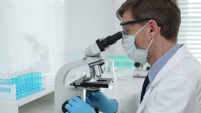 Masked male scientist in lab coat, protective glasses and blue gloves is adding reagent to test tube with microscopes and test tube rack in modern laboratory. Medicine, healthcare and science concept