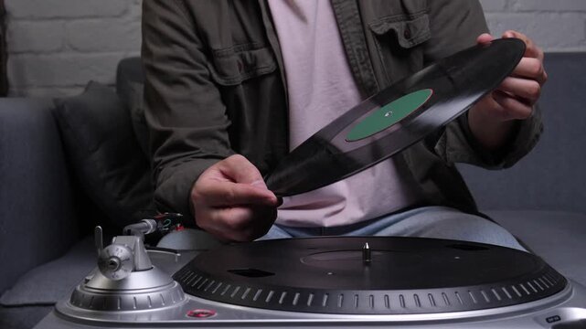 Person's hands preparing to place a black vinyl record with a green label onto a modern silver turntable, engaging with music through an analog and vintage audio experience