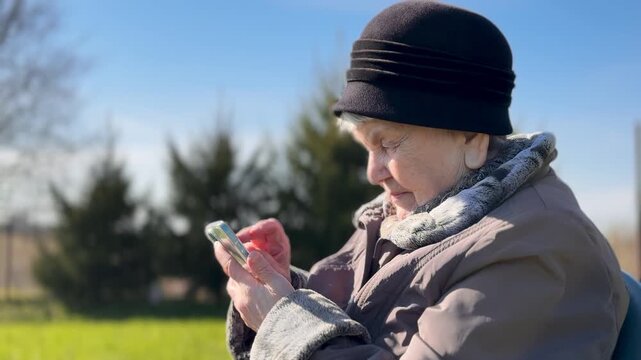 Elderly grandmother reading news on smartphone, seated on chair in sunlit garden plot, bundled coat and knit hat, scrolling