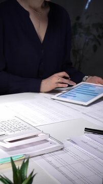 Woman working late night in office, multitasking with a tablet and writing on documents, managing heavy paperwork load. Audit and taxes in business