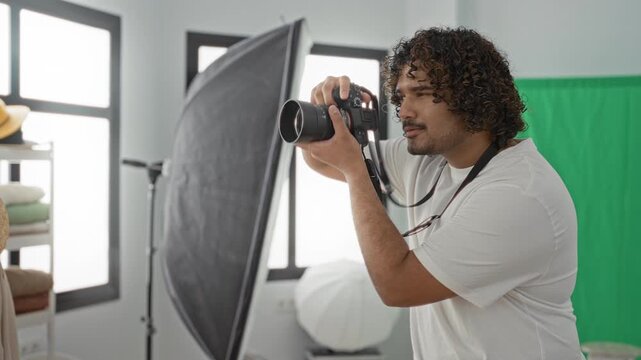 Young indian man holds dslr to eye, adjusts lens and camera dial, waves hand while smiling beside green screen and softbox in studio; concentration creativity.