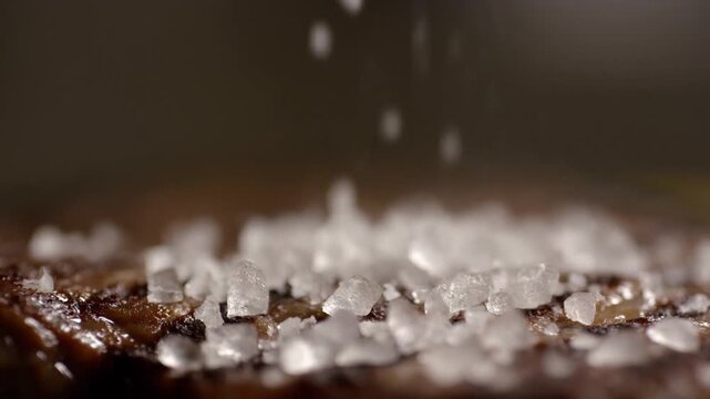 Slow motion close up of coarse salt crystals sprinkling onto grilled steak surface on kitchen countertop for seasoning shot
