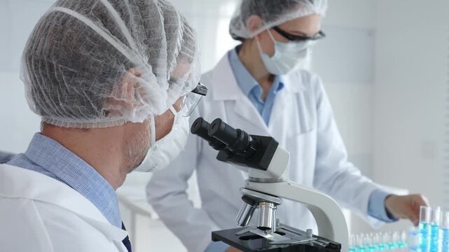 Senior male scientist wearing protective mask, glasses and hairnet using microscope with young female colleague assisting, holding tablet and taking notes in laboratory. Medicine and science concept