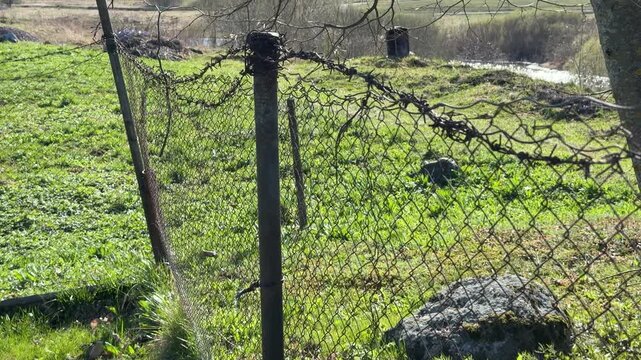 Rusty chain fence on garden plot under bare tree branches, leaning metal post and old pipe casting long shadow across fresh spring.