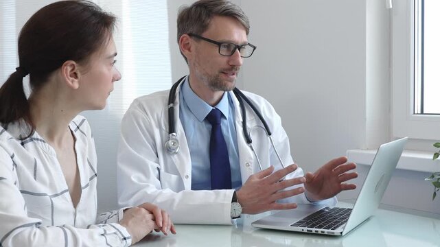 Male doctor showing medical test results on a laptop to a female patient, discussing diagnosis and treatment options in a bright medical office. Medicine and health care