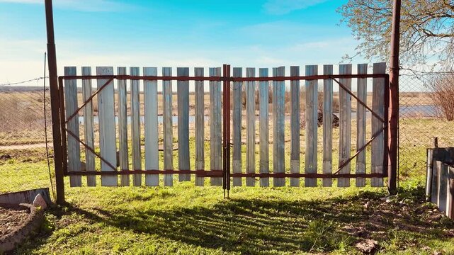 Weathered metal gate with vertical stainless strips, chain link fences flanking sides, spring grass carpeting ground, clear blue