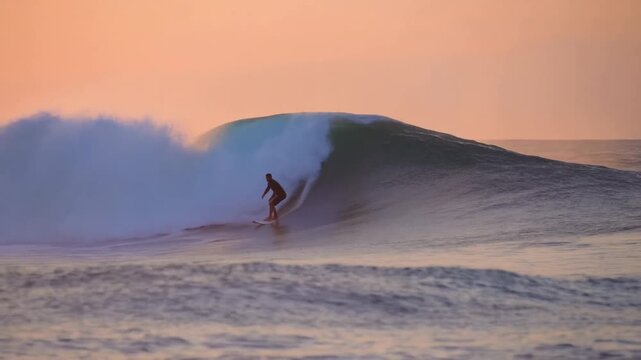 Tracking shot of surfer riding barreling wave at sunrise over reef break on calm ocean near shore