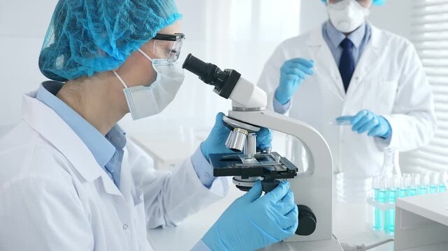 Female scientist wearing protective gear is examining specimen under microscope while man laboratory assistant handles glassware in a professional medical investigation. Medicine and science concept