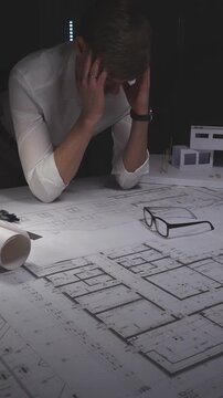Architect feeling stress and burnout while working late on building blueprints and construction plans with a headache. Concept of architecture and business in low key lighting