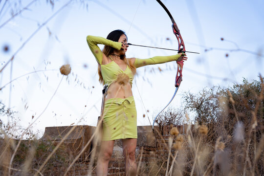 Asian woman practicing archery, focusing with a recurve bow and arrow, showing skill and determination outdoors